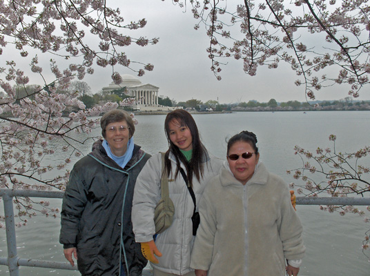Dona, Ann and Nanta stand in front of the railing with water behind them.  Cherry tree blossoms hang over behind them, and on the other side of the water is the Jefferson Memorial.